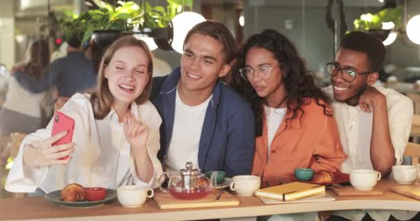 Jeunes gens riant et saluant tout en posant pour selfie dans un restaurant confortable. Fille heureuse tenant smartphone horizontalement tout en prenant des photos avec des amis. Concept d'émotions positives. — Video