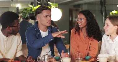 Modern kafede iş arkadaşlarına yemek anlatırken konuşan ve el kol hareketi yapan yakışıklı bir adam. Çeşitli gençler gülümserken erkek arkadaşları dinliyorlar. İletişim kavramı.