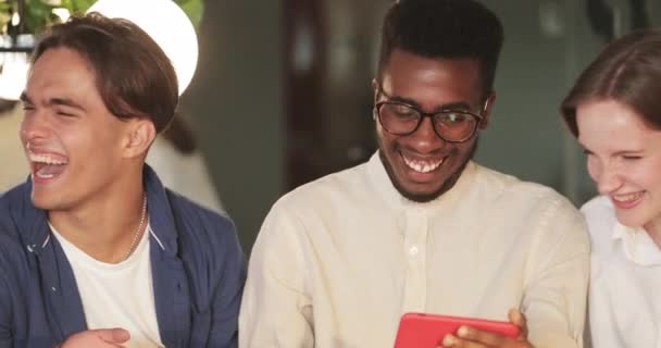 Blisko afro amerykański facet pokazuje swoim przyjaciołom zabawne wideo, gdy siedzą w przytulnej kawiarni. Szczęśliwi przyjaciele śmieją się i patrząc na ekran telefonu komórkowego podczas spędzania czasu razem. — Wideo stockowe