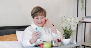 Gülümseyen, el sallayan ve konuşan neşeli yaşlı bayan akıllı telefonunu iletişim için kullanıyor. Evde yaşayan mutlu yaşlı kadın Rob yatakta kahvaltı ederken görüntülü konuşma yapıyor..
