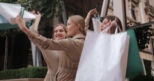 Üç mutlu kız selfie çekiyor ve sokakta dikilirken alışveriş torbalarını kaldırıyor. Güzel milenyum bayan arkadaşları gülümsüyor ve poz veriyor telefon kullanıyor ve ekrana bakıyor.. — Stok video