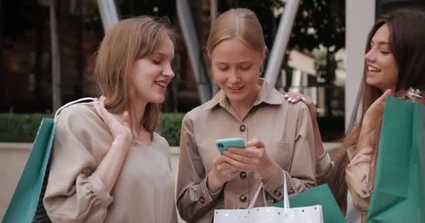 Belles femmes tenant des sacs à provisions riant tout en se tenant à la rue. Attrayant millénial fille en utilisant smartphone tandis que ses amies regardent l'écran, parler et rire. — Video