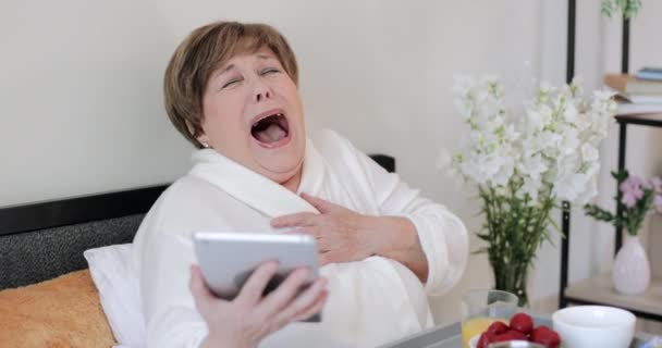 Mujer mayor en casa roba riendo mientras mira la pantalla de su gadget moderno. Anciana jubilada de buen humor viendo videos divertidos mientras usa la tableta y desayunando en la cama . — Vídeo de stock