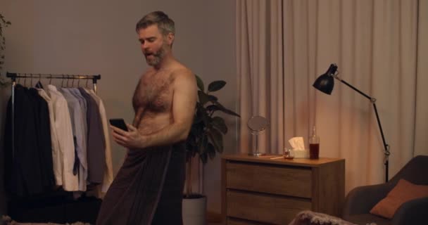 Przystojniak po czterdziestce udający, że gra na gitarze i śpiewa w domu. Dojrzały pogodny mężczyzna w ręczniku kąpielowym bawiący się patrząc na smartfona i pisząc na maszynie. Pojęcie czasu wolnego. — Wideo stockowe