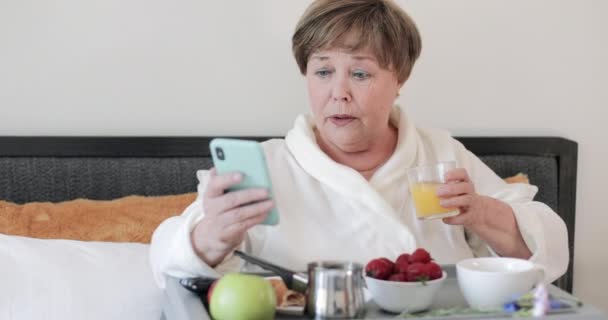 Oudere mooie dame het maken van geschokte gezichtsuitzetting tijdens het gebruik van haar smartphone. volwassen vrouw in huis rob het hebben van ontbijt en drinken sap terwijl het krijgen van bed nieuws op haar telefoon. — Stockvideo