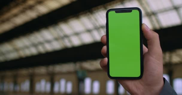 Крупним планом чоловіча рука тримає порожній зелений екран мобільного телефону на залізничній станції. Концепція хромового ключа, сміх і технологія. Розмитий фон . — стокове відео