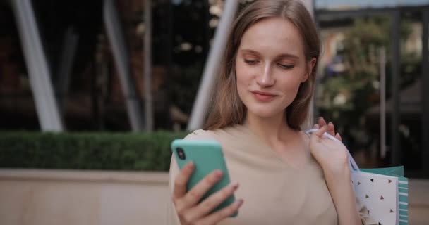 Vista de cerca de la chica bonita usando su teléfono inteligente mientras está de pie en la calle. Hermosa mujer milenaria sosteniendo bolsa de papel de compras y sonriendo mientras se comunica en las redes sociales . — Vídeos de Stock