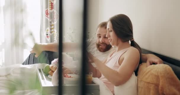 Pareja feliz acostada en la cama, riendo y viendo fotos divertidas en el teléfono inteligente. Hombre y mujer de buen aspecto desayunando, y hablando mientras mira la pantalla en el dormitorio . — Vídeos de Stock