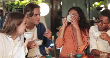 Rahat iş arkadaşları çay içiyor, gülüyor ve rahat bir restoranda otururken el kol hareketi yapıyor. Gülümseyen genç insanlar birlikte iyi vakit geçirirken pozitif duygular gösteriyorlar..