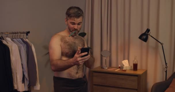 Pohledný dospělý chlap dostává dobré zprávy na smartphone a radovat se při čištění zubů. Vousatý muž v osušky dostat zprávu a začít tancovat doma.. — Stock video
