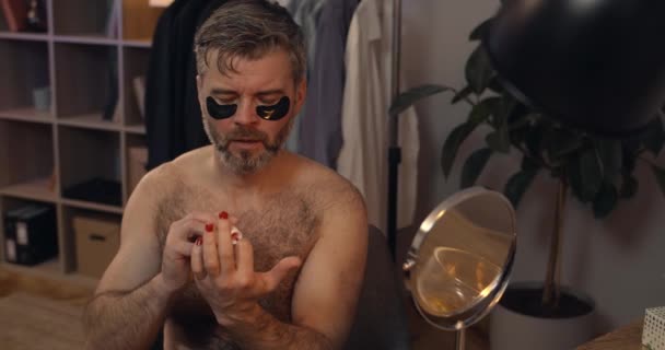 Brutal mec mature peignant des ongles avec du vernis rouge et la tête tremblante. Homme barbu dans les années 40 avec des patchs pour les yeux faisant une procédure de beauté tout en étant assis devant le miroir. Concept de beauté. — Video