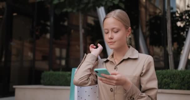 Close up widok atrakcyjnej blondynki włosy za pomocą smartfona podczas spaceru. Tysiąclecia kobieta patrzy na ekran telefonu podczas zakładania i noszenia papierowej torby na ramię. — Wideo stockowe