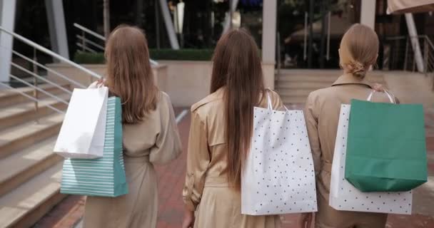 Vue arrière de jeunes femmes élégantes marchant et portant des sacs en papier après les achats. Millennial amies tournant la tête, souriant et regardant la caméra à la rue de la ville. — Video