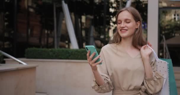 Close-up zicht van millennial vrolijk meisje met behulp van haar smartphone tijdens het lopen op straat. mooie vrouw kijken naar telefoon scherm en glimlachen terwijl het dragen van papieren zak op de schouder. — Stockvideo