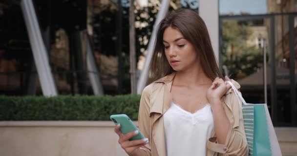 Mujer morena bonita usando su teléfono móvil y mirando a la cámara mientras está de pie en la calle. Retrato de chica alegre navegando por Internet mientras sostiene la bolsa de papel de compras y sonriendo . — Vídeo de stock