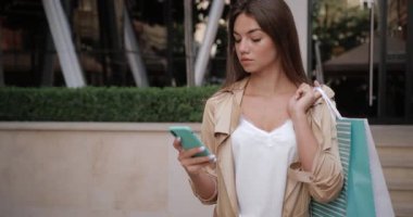 Kameraya bakıp cep telefonunu kullanırken gülümseyen esmer kızın yakın görüntüsü. Sokakta omzunda alışveriş poşeti olan çekici bir kadının portresi..