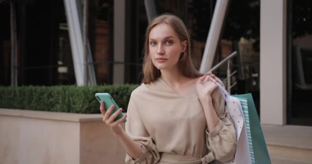 Mujer atractiva levantando la cabeza y mirando a la cámara mientras navega por Internet en el teléfono inteligente. Retrato de una chica bonita sosteniendo una bolsa de papel en el hombro mientras está de pie en la calle de la ciudad. Acercar . — Vídeos de Stock