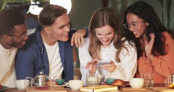 Jolie fille millénaire tenant smartphone tout en montrant des photos d'amis. Groupe de personnes multiethniques riant et regardant l'écran du téléphone tout en passant du bon temps ensemble dans un café. — Video