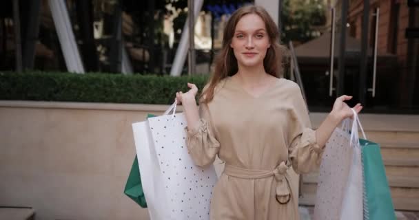 Jovencita alegre caminando y dando la vuelta con bolsas de papel. Atractiva chica millennial mirando a la cámara y sonriendo mientras lleva sus compras después de ir de compras en la calle de la ciudad . — Vídeo de stock