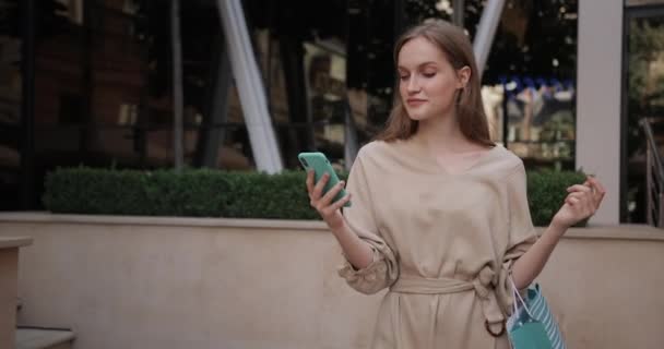 Вигляд на стильну молоду жінку, яка використовує свій смартфон під час ходьби на вулиці. Гарна дівчина, яка носить сумку з паперу для покупок і посміхається під час балачки в соціальних мережах . — стокове відео