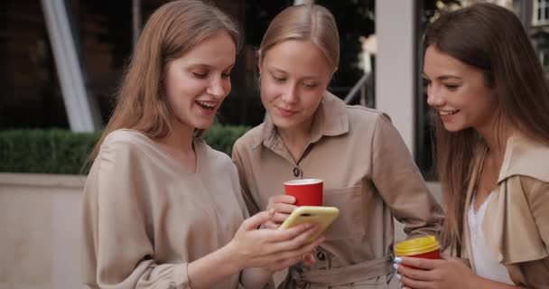 Привлекательные девушки держат бумажные кофейные чашки и улыбаются, стоя на улице. Молодая женщина использует смартфон в то время как ее подруги смотрят на экран, разговаривают и смеются. — стоковое видео