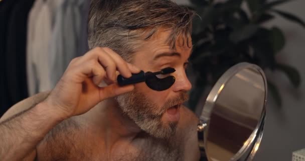 Brutal mature guy applying black lashes mascara and shaking head. Bearded handsome man in 40s with eye patches sitting in front of mirror at home in evening. Concept of beauty. — Stock Video