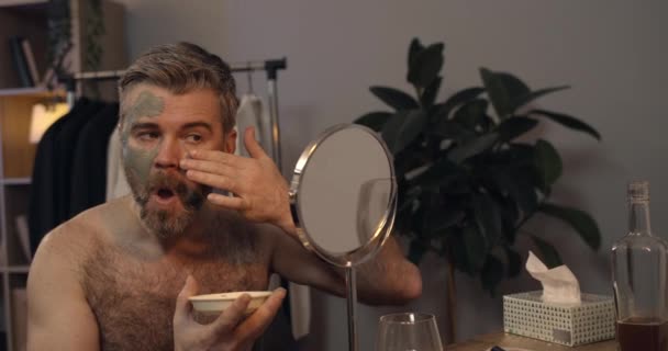 Vue de la récolte d'un bel homme joyeux mettant masque facial purifiant et sentant. Brutal homme barbu dans les années 40 regardant dans le miroir tout en faisant la procédure de beauté à la maison. Concept de beauté. — Video