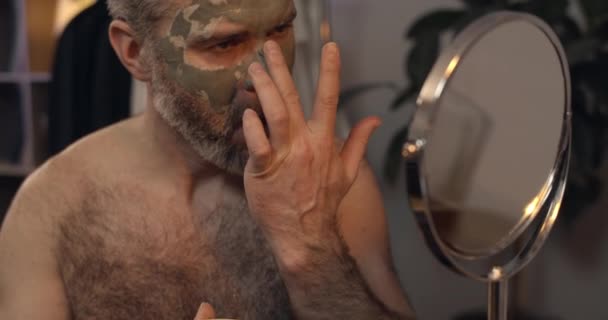 Közelről láthatjuk, ahogy a szakállas, érett fickó befejezi az arcpakolást és rázza a fejét. Vonzó férfi a 40-es éveiben, aki kozmetikázik és tükörbe néz. A szépség és a gondozás fogalma. — Stock videók