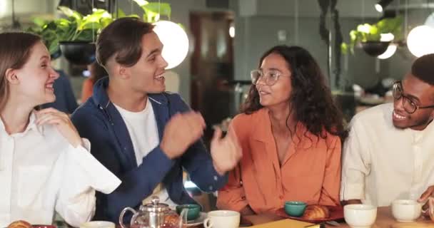 Millennial emotionaler Mann gestikuliert und redet, während er mit multiethnischen Mitarbeitern in einem gemütlichen Restaurant sitzt. Glückliche junge Leute, die zuhören und lachen. Kommunikationskonzept. — Stockvideo