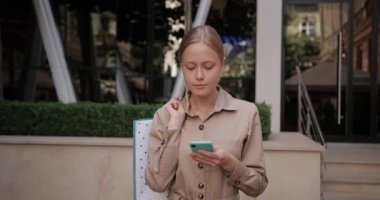 Sarışın güzel bir kadının yürürken akıllı telefonunu kullanmasının görüntüsü. Millennial çekici kız telefon ekranına bakarken omzunda alışveriş poşeti taşıyor..
