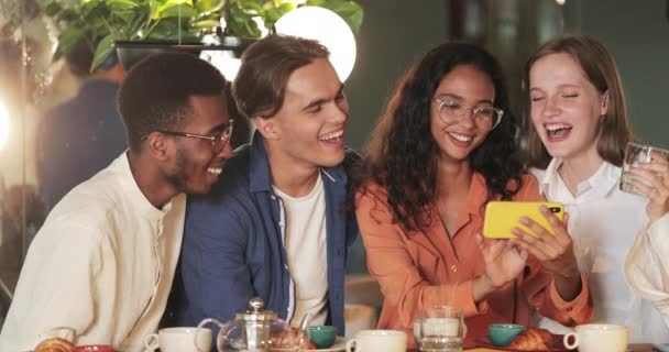 Attrayant fille tenant le téléphone horizontalement tout en montrant des amis des photos drôles. Millennial personnes gaies riant et regardant l'écran mobile tout en étant assis dans un café confortable. — Video