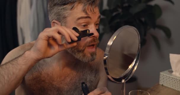 Beau homme mature appliquant mascara avec bouche ouverte tout en étant assis devant le miroir.Homme barbu avec des patchs oculaires se préparer pour la date dans la soirée. Accueil arrière-plan. — Video