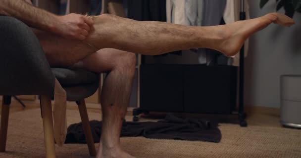 Schnittansicht einer männlichen Person mit nacktem Oberkörper, die einen Strumpf anzieht, während sie auf einem Stuhl sitzt. Hintergrund Heimat. — Stockvideo