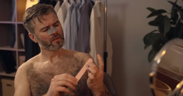 Close up view of mature guy filing nails while sitting at chair in front of mirror. Bearded man in 40s with nose pore cleansing strip doing beauty procedure at home in evening. — Stock Video