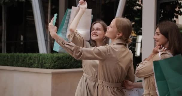 Wesołe dziewczyny trzymając torby na zakupy podczas robienia selfie i patrząc na ekranie smartfona. Atrakcyjne kobiety uśmiechające się i pozujące podczas stania na ulicy i korzystania z telefonu. — Wideo stockowe