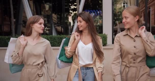 Millenial pretty girls with paper bags communicating while walking at city street. Female attractive friends talking and smiling while discussing their shopping. Concept of friendship. — Stock Video