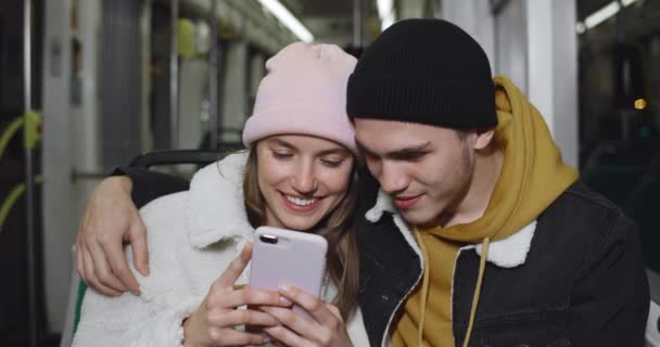 Вид на тисячолітню пару, дивлячись на екран смартфона, дивлячись відео та посміхаючись. Приваблива дівчина тримає телефон, сидячи зі своїм хлопцем на сидіннях у громадському транспорті . — стокове відео