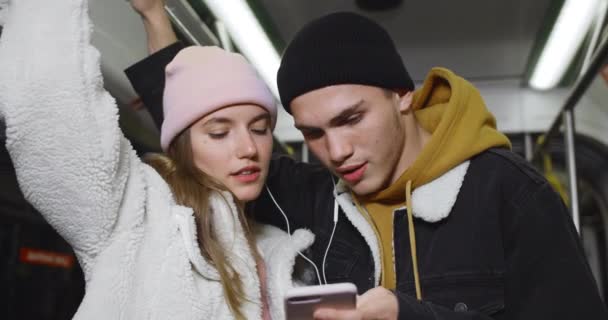 Vista de cerca de la feliz pareja riendo mientras comparte auriculares. Millennial chico y chica usando el teléfono y mirando a la pantalla mientras van en transporte público por la noche . — Vídeos de Stock