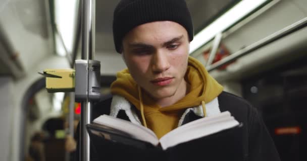 Millennial man het lezen van interessante boek tijdens het gaan in het openbaar vervoer. Jonge knappe man met een draadloze koptelefoon die geniet van het verhaal terwijl hij 's avonds naar huis pendelt. Begrip levensstijl. — Stockvideo