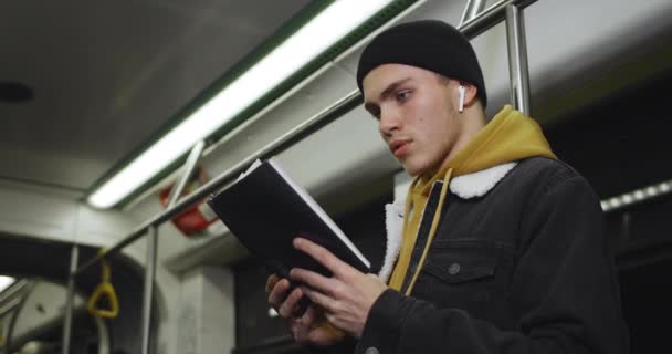 Vue latérale du jeune homme avec des écouteurs sans fil tournant des pages tout en enjolivant l'histoire. Millennial beau garçon lisant livre tout en allant dans les transports publics en soirée. Concept de vie réelle. — Video