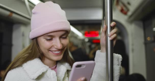 Joven alegre en sombrero rosa sonriendo mientras usa su teléfono inteligente y va en transporte público. Millennial chica guapa leyendo un mensaje divertido mientras chatea en las redes sociales . — Vídeo de stock