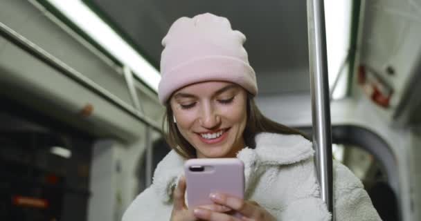 Chica bonita en auriculares inalámbricos utilizando el teléfono inteligente y sonriendo mientras viaja en transporte público. Millennial buen aspecto en la pantalla del teléfono mientras escribe el mensaje en las redes sociales . — Vídeos de Stock