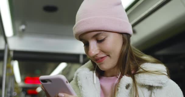 Nahaufnahme einer hübschen Millennial-Frau, die ihr Smartphone benutzt, während sie in öffentlichen Verkehrsmitteln unterwegs ist.Attraktives Mädchen mit Kopfhörern, das lächelt und beim Chatten in den sozialen Medien auf den Handybildschirm schaut. — Stockvideo