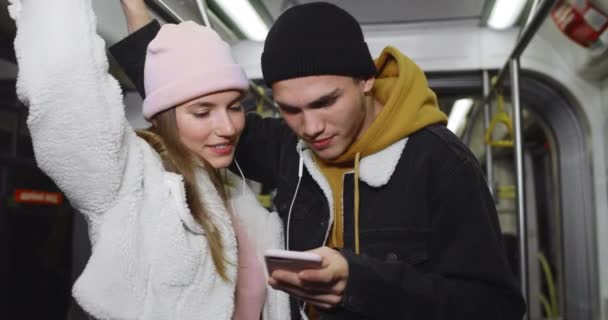 千禧年夫妇聊天，看手机屏幕。漂亮的女孩和她的男朋友在晚上乘坐公共交通工具时共用耳机，笑着. — 图库视频影像