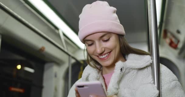 Joven mujer alegre en sombrero rosa navegando por Internet y sonriendo en metro. Milenial chica guapa comunicándose en las redes sociales mientras va en transporte público . — Vídeo de stock