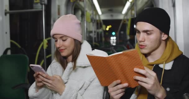 Millennial τύπος χρησιμοποιώντας το βιβλίο ανάγνωσης, ενώ κάθεται κοντά στην υπέροχη φίλη του. Νέα όμορφη κοπέλα που χρησιμοποιεί smartphone ενώ πηγαίνει σε δημόσιες συγκοινωνίες με το φίλο της. Έννοια της πραγματικής ζωής. — Αρχείο Βίντεο
