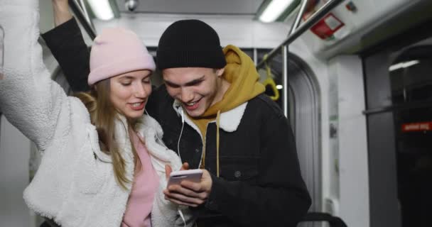 Jeune homme et sa petite amie en utilisant smartphone et rire tout en regardant l'écran.Couple joyeux partage écouteurs et regarder vidéo drôle tout en allant dans les transports en commun. — Video