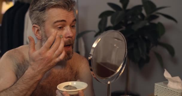 Mature beau homme appliquant masque facial tout en étant assis devant le miroir. Barbu gars dans les années 40 faisant beauté prosedure dans la soirée à la maison. Concept de soin et de beauté. — Video