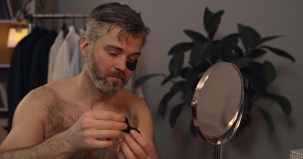 Vue rapprochée d'un bel homme dans les années 40 appliquant des patchs pour les yeux noirs et regardant dans le miroir.Mature célibataire barbu faisant procédure de beauté tout en préparant à ce jour à son appartement. — Video