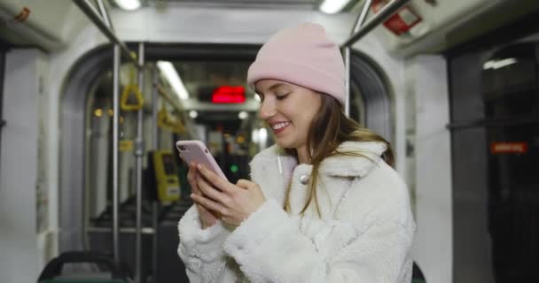 Jovencita alegre mirando la pantalla de su smartphone y sonriendo. Vista de la cosecha de chica atractiva en los auriculares usando el teléfono mientras va en transporte público. Concepto de la vida real. — Vídeo de stock